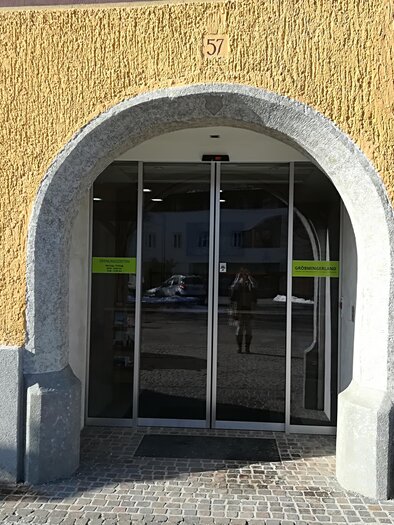 An entrance area with a glass sliding door and an arched façade. On the left, there is a green information sign with a symbol. | © TVB Gröbminger Land 