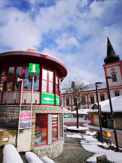 Ein Informationsstand in einer verschneiten Stadt. Im Hintergrund sieht man eine Kirche mit einem hohen Turm. | © TV Hochsteiermark