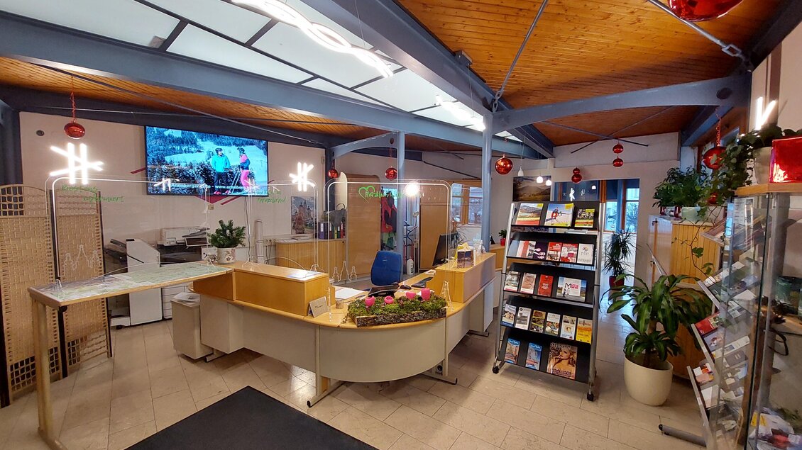 Ein helles Empfangsgebiet mit einem modernen Empfangstresen und einer Bücherwand. Im Hintergrund ist ein Bildschirm zu sehen, das Ambiente ist freundlich und einladend. | © TV Hochsteiermark