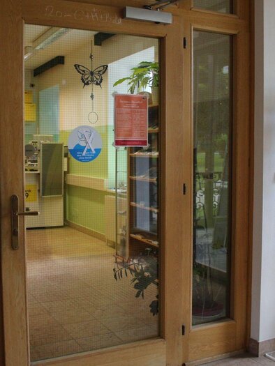An inviting entrance to a room with bright walls and plants. In the entrance area, there are information signs and a brochure stand. | © Viola Lechner