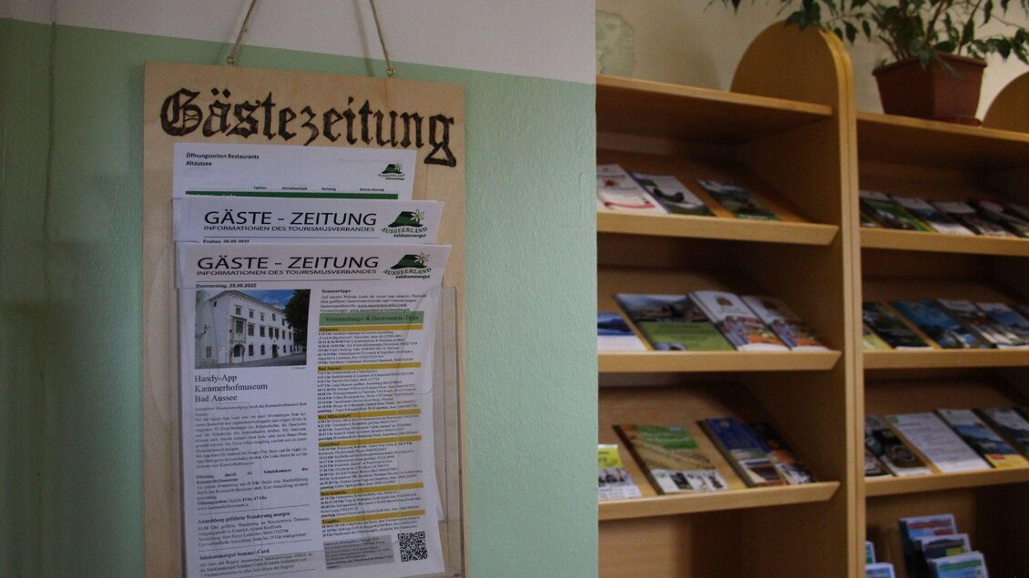 Ein Informationsbrett mit mehreren Exemplaren der "Gästezeitung" hängt an der Wand. Im Hintergrund sind Regale mit verschiedenen Broschüren und Informationsmaterialien zu sehen. | © Viola Lechner