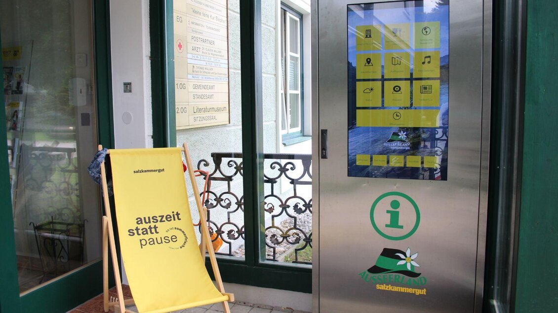 Ein gelbes Liegestuhl mit der Aufschrift „auszeit statt pause“ steht vor einer Informationsstelle. Die Eingangstür ist aus Glas und hat ein digitales Display. | © Viola Lechner