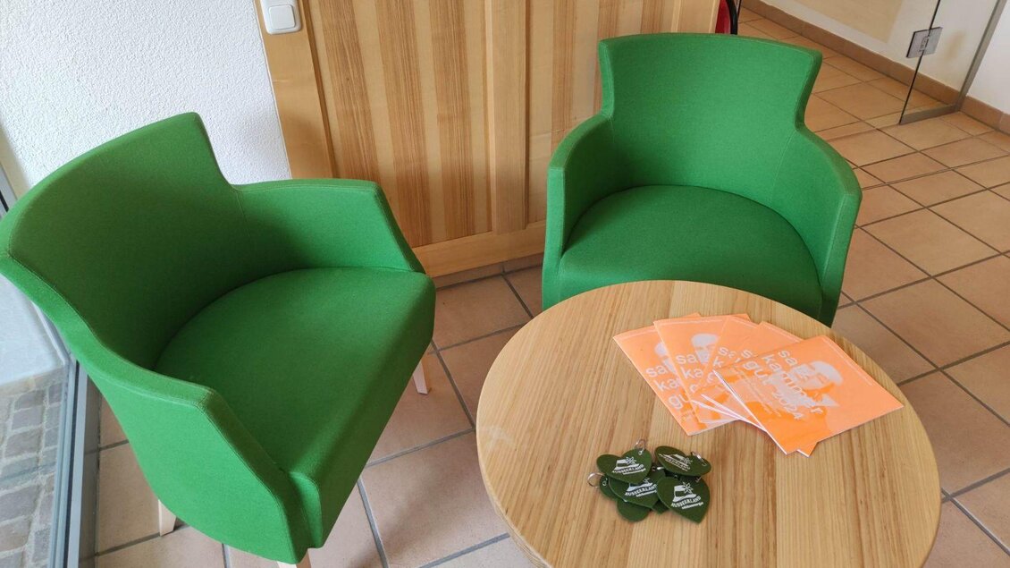 Zwei grüne Stühle stehen an einem kleinen Tisch. Auf dem Tisch liegen orangefarbene Flyer und einige Schlüsselanhänger. | © Johanna Provatopoulos