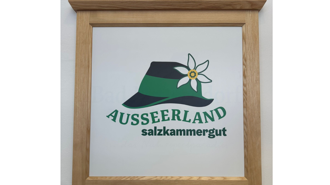 Ein dekoratives Schild mit einer stilisierten Hutgrafik und einer Blume. Darunter steht der Schriftzug "Ausseerland Salzkammergut". | © Daniela Casari