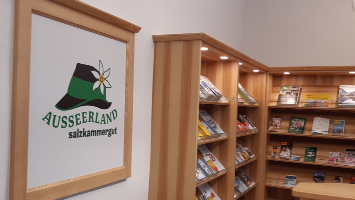 Ein Informationsbereich mit einer Holzregalwand und verschiedenen Broschüren. Im Hintergrund ist ein Logo mit dem Schriftzug "Ausseerland Salzkammergut" zu sehen. | © Daniela Casari