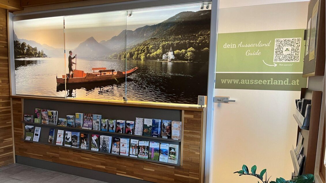 Ein heller, einladender Empfangsbereich mit einem großen Bild eines Sees und Berge im Hintergrund. Darunter befindet sich ein Regal mit Informationsmaterialien über das Ausseerland. | © Sophie Eggenreitter