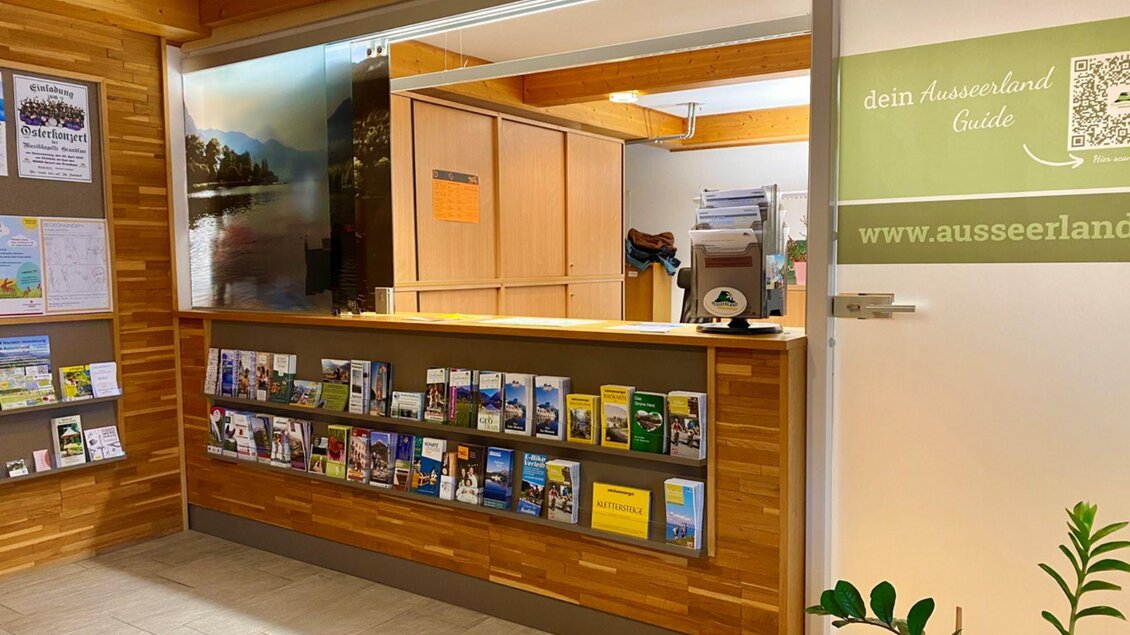Ein Empfangsbereich mit einem Holztresen und einem Regal voller Informationsbroschüren. Im Hintergrund sind einige Büromöbel und eine Wand mit Bildern der Natur zu sehen. | © Bettina Scheck