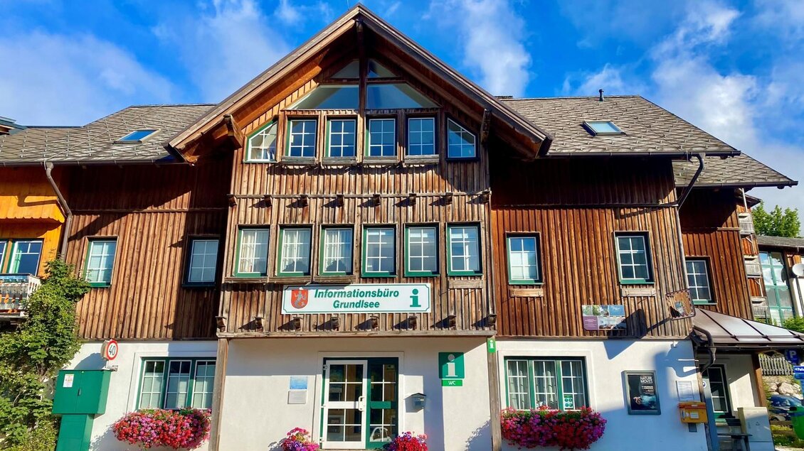 Ein traditionelles Holzgebäude mit mehreren Fenstern und einem Informationsbüro. Vor dem Gebäude blühen bunte Blumen. | © Bettina Scheck