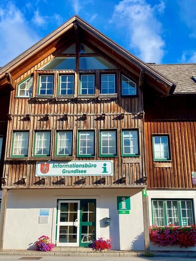A traditional wooden building with several windows and an information office. Colorful flowers are blooming in front of the building. | © Bettina Scheck