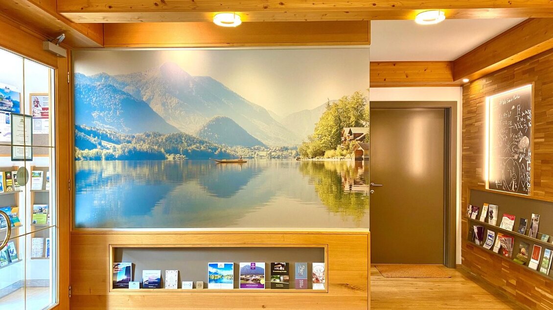 Ein einladender Empfangsbereich mit Holzverkleidung. An der Wand hängt ein großes Landschaftsbild eines ruhigen Sees, umgeben von Bergen. | © Bettina Scheck