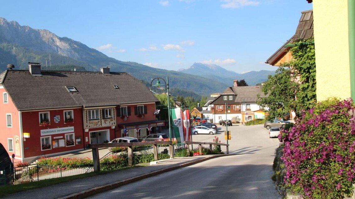 Eine malerische Straße mit mehreren Gebäuden und einem Blick auf die Berge. Die Umgebung ist grün und einladend, mit Blumen am Rand. | © TVB Ausseerland Salzkammergut/Kolb