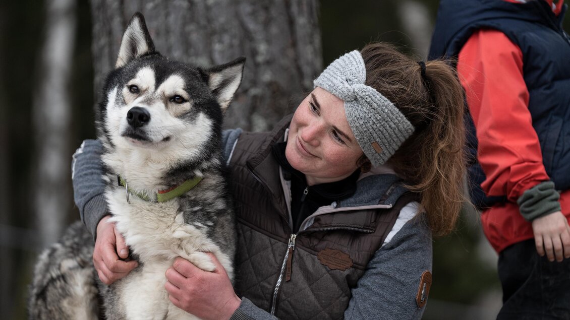 Eine Frau umarmt einen Hund in der Natur. Im Hintergrund sind weitere Personen und Bäume zu sehen. | © Harald Tauderer