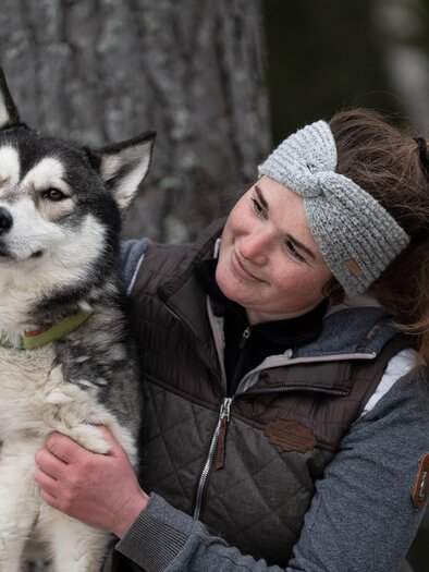 A woman is hugging a dog in nature. In the background, other people and trees can be seen. | © Harald Tauderer