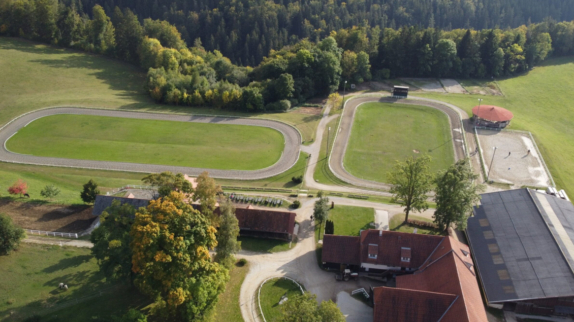 Eine weitläufige Rennbahn mit zwei Bahnen, umgeben von grünen Wiesen und Bäumen. Im Vordergrund sind einige Gebäude und eine farbenfrohe Landschaft zu sehen. | © Piet Hoyos-David Schabl