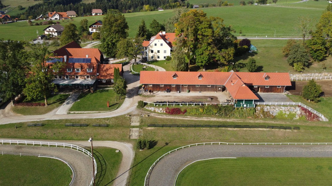 Eine Übersicht eines schönen Reiterhofs mit mehreren Gebäuden und grünen Wiesen. Im Hintergrund sind sanfte Hügel und Bäume zu sehen. | © Piet Hoyos-David Schabl
