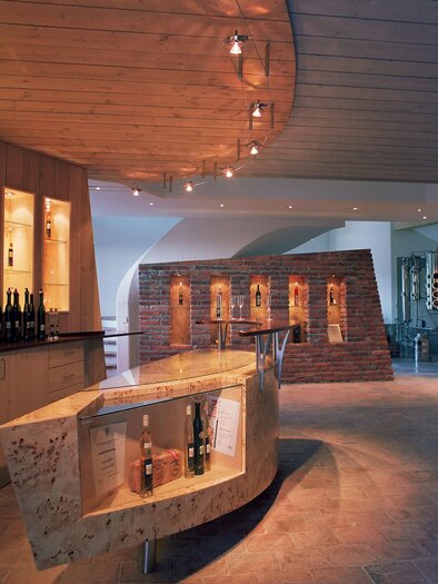 A modern wine tasting room with a curved bar. In the background are wine barrels and a stylish wall with bottles. | © Brennerei Jöbstl