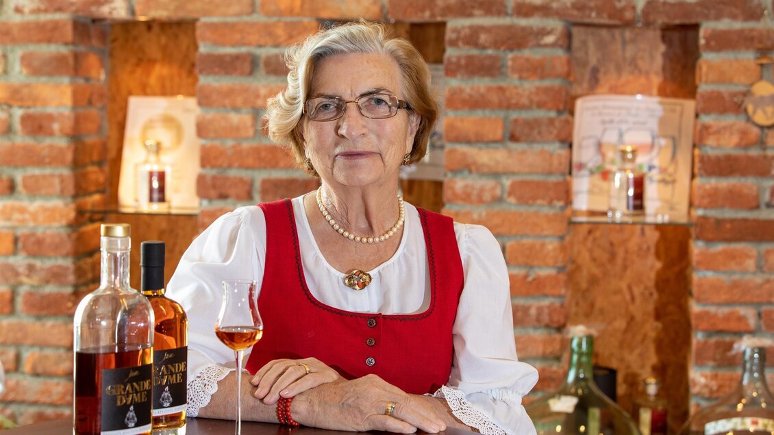 Eine ältere Dame in traditioneller Tracht, die an einem Holztisch sitzt. Neben ihr stehen zwei Flaschen Schnaps und ein Glas mit einem Getränk. | © Brennerei Jöbstl