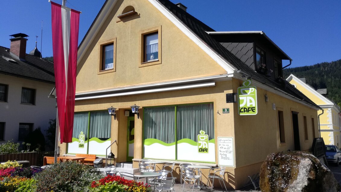 Ein gemütliches Café mit einer gelben Fassade und großen Fenstern. Vor dem Café stehen Tische und Stühle umgeben von bunten Blumen.