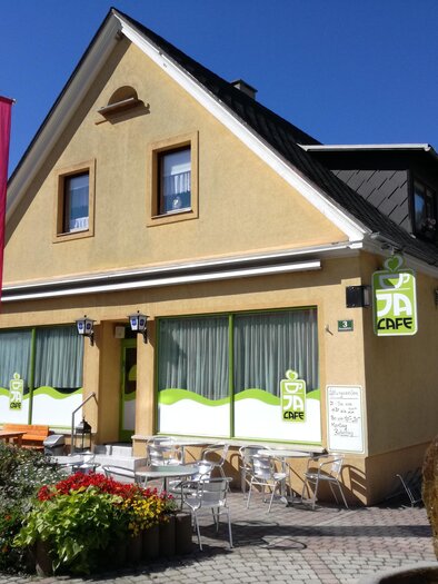 A cozy café with a yellow facade and large windows.  
In front of the café are tables and chairs surrounded by colorful flowers.