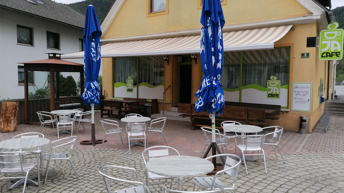 Ein gemütliches Café mit einer Terrasse und weißen Tischen. Blaue Sonnenschirme bieten Schatten in der Außenanordnung.