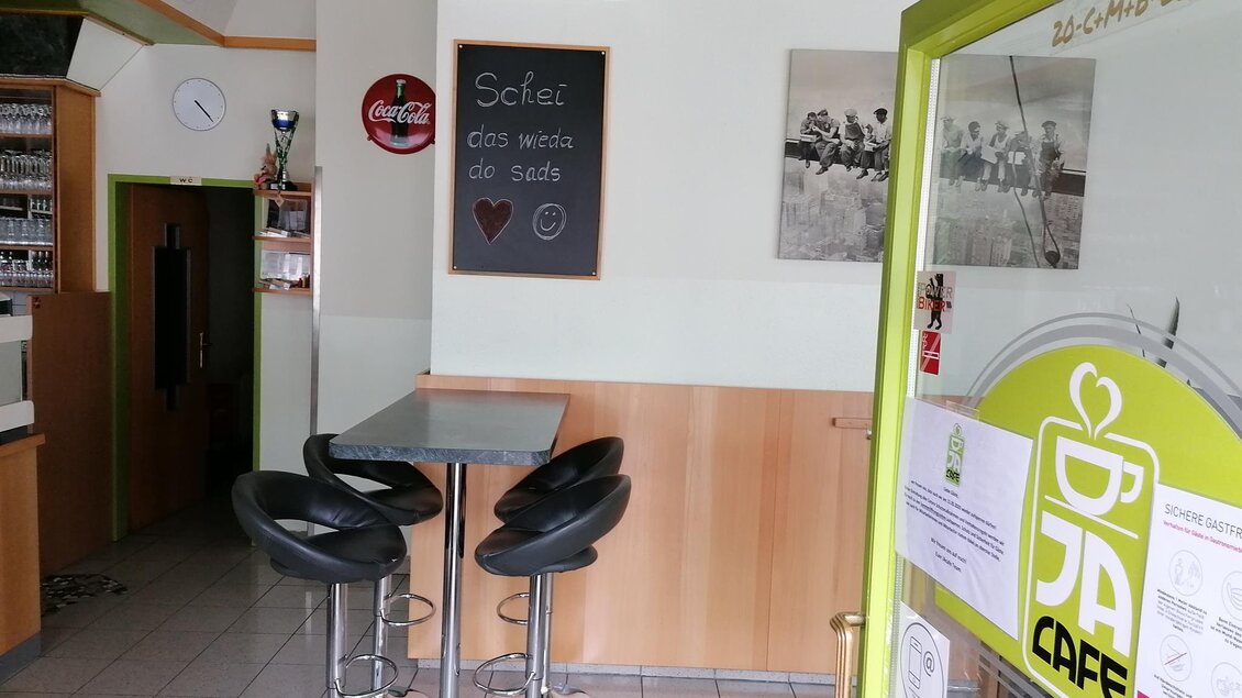 Ein kleiner, einladender Café-Bereich mit hohen Stühlen und einem Tisch. An der Wand hängt eine Tafel mit einer herzlichen Botschaft.