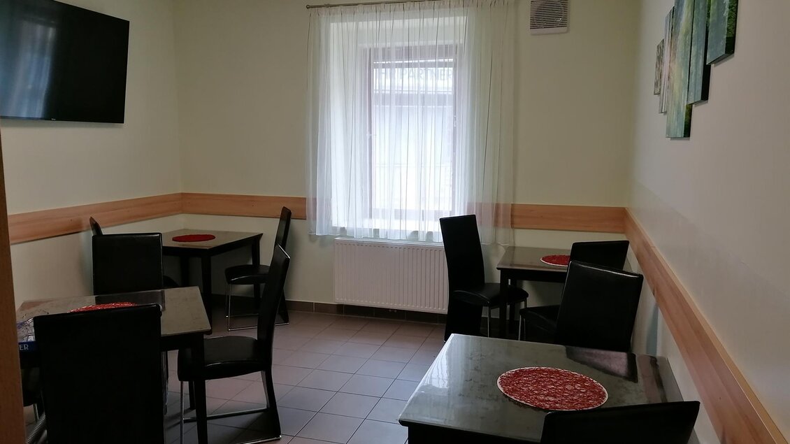 Ein helles, gemütliches Zimmer mit Tischen und Stühlen. An der Wand hängt ein Fernseher, und es gibt Fenster mit Vorhängen.