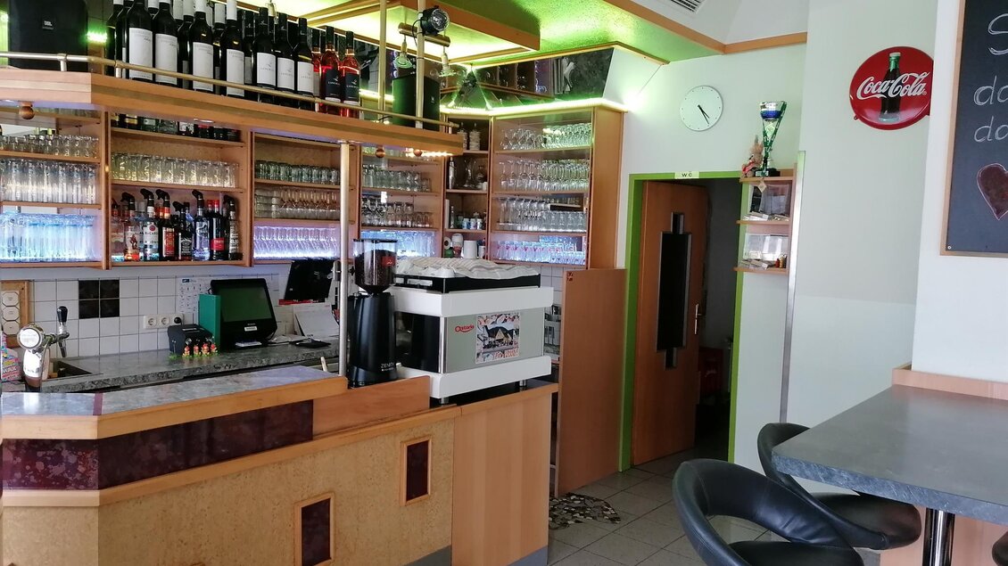 Eine gemütliche Bar mit einem Bartresen, Regalen voller Flaschen und einem modernen Kaffeeautomaten. Es gibt einige Barhocker und eine ruhige Atmosphäre.