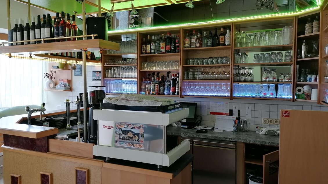 Eine gemütliche Bar mit einer Holztheke und Regalen voller Getränke. Die Atmosphäre wird durch grüne Beleuchtung aufgepeppt.