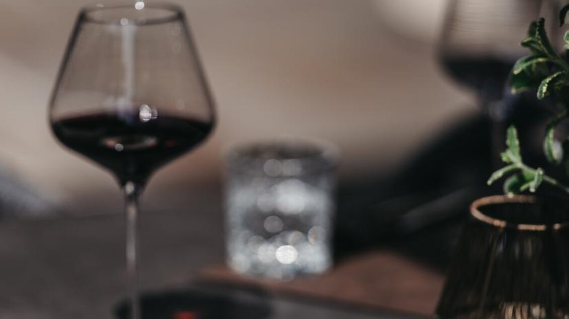 Ein elegantes Gericht mit Fleisch und buntem Gemüse auf einem weißen Teller. Daneben steht ein Glas Rotwein und eine stilvolle Tischdekoration. | © JOHANN Schladming