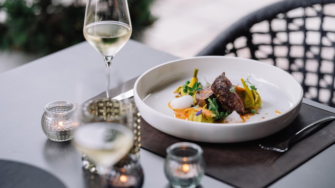 Ein elegantes Gericht auf einem Tisch, umgeben von Gläsern und Kerzenlicht. Das Gericht besteht aus Fleisch, Gemüse und einer kreativen Präsentation. | © JOHANN Schladming