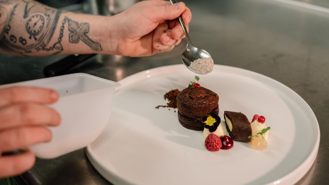 Ein Koch verziert ein Dessert auf einem weißen Teller. Das Dessert besteht aus Schokoladenkuchen, frischen Beeren und einer Creme. | © JOHANN Schladming