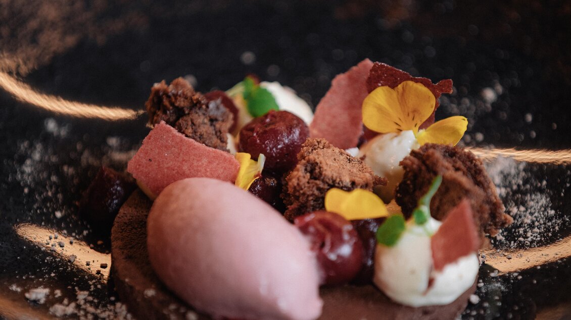 Ein kunstvoll angerichtetes Dessert auf einem dunklen Teller. Es besteht aus Schokoladenkuchen, Sahne, Kirschen und einem Himbeersorbet, garniert mit essbaren Blüten. | © JOHANN Schladming
