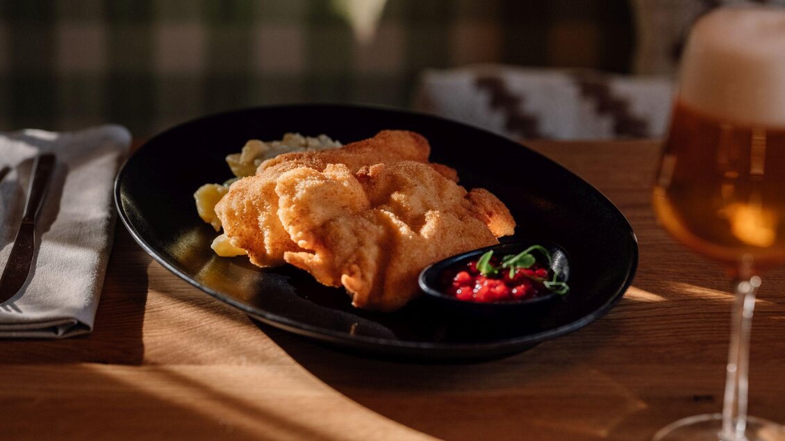Ein Teller mit knusprigem Fisch und Beilage, serviert mit einer kleinen Schale Sauce. Im Hintergrund steht ein Glas Bier. | © JOHANN Schladming