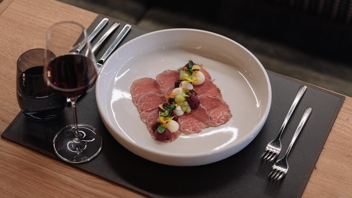 Ein elegantes Gericht mit dünn geschnittenem Fleisch, garniert mit frischen Kräutern und Gemüse. Dazu ein Glas Rotwein auf einem stilvollen Tisch. | © JOHANN Schladming