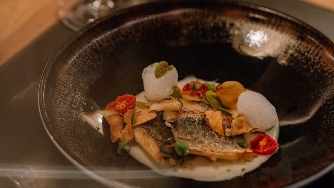 Ein kunstvoll angerichteter Teller mit Fisch, buntem Gemüse und kleinen Beilagen. Im Hintergrund steht ein elegantes Weinglas auf einem Holztisch. | © JOHANN Schladming
