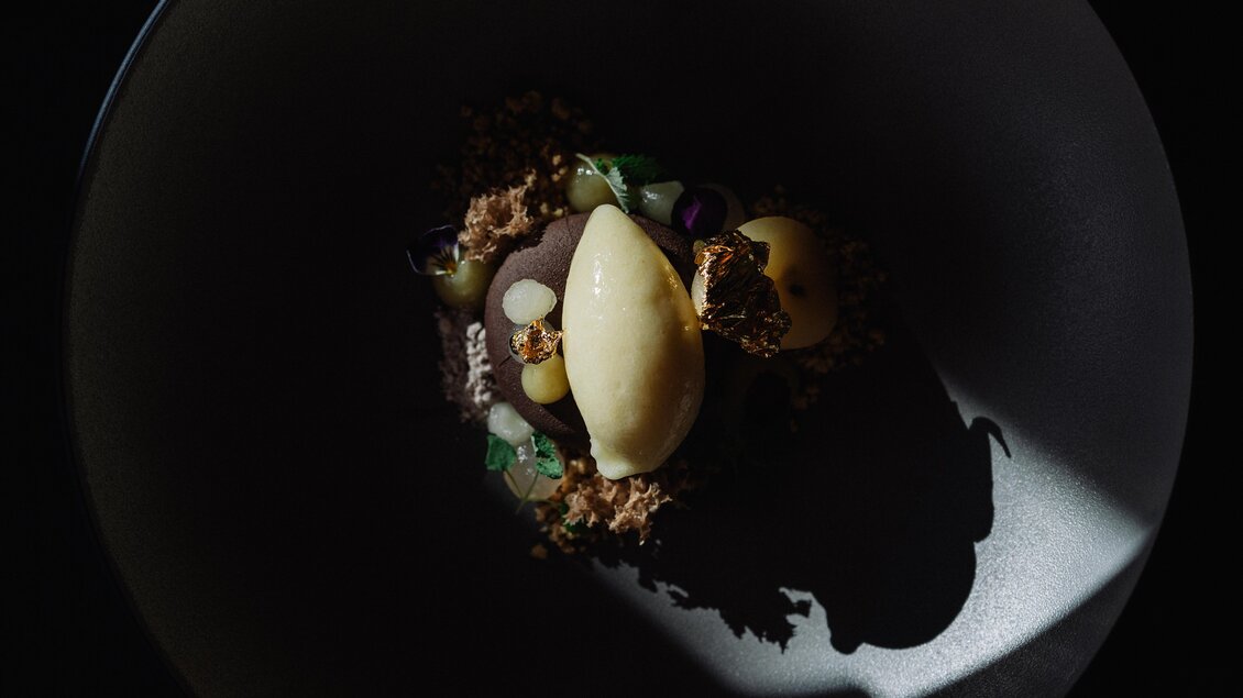 Ein kunstvoll angerichtetes Dessert in einer dunklen Schale. Es besteht aus einer cremigen Sorbetkugel, frischen Früchten und knusprigen Elementen. | © JOHANN Schladming