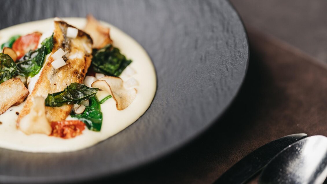 Ein delikates Gericht auf einem schwarzen Teller, bestehend aus gebratenem Fisch, Gemüse und einer cremigen Sauce. Frische Spinatblätter und Tomaten verleihen dem Gericht Farbe und Geschmack. | © JOHANN Schladming