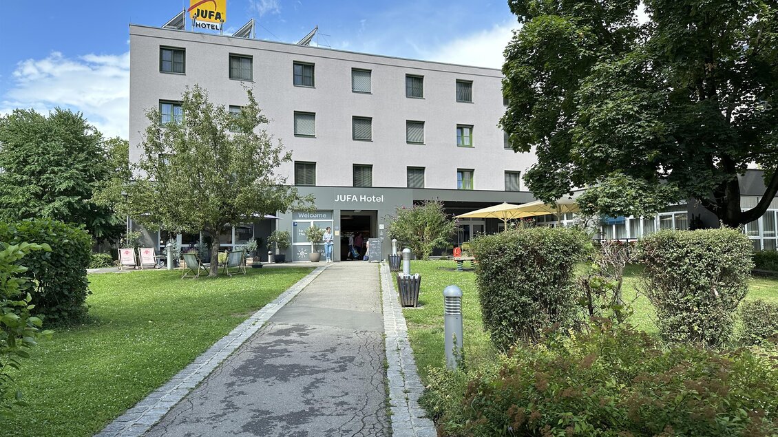 Ein modernes Hotel mit grünem Garten und angenehmem Eingangsbereich. Über dem Gebäude ist ein gelbes Schild mit der Aufschrift "JOFA". | © JUFA Hotels