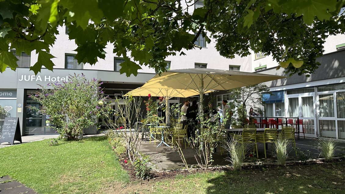 Ein Außenbereich des JUFA Hotels mit einem Sonnenschirm und Sitzgelegenheiten im grünen Garten. Einige Gäste genießen die entspannte Atmosphäre. | © JUFA Hotels