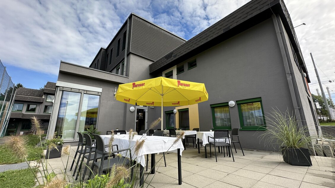 Eine moderne Terrasse mit Tischen und Stühlen unter einem gelben Sonnenschirm. Im Hintergrund ist ein elegantes Gebäude und ein klarer Himmel zu sehen. | © JUFA Hotels