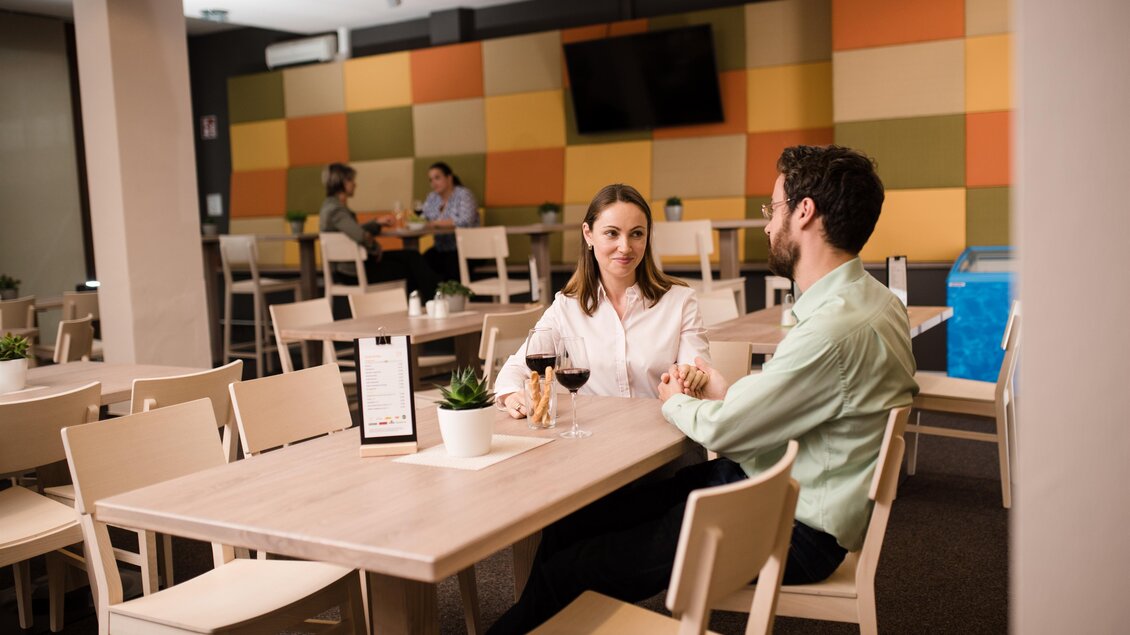 Gäste mit Getränken an Holztischen im hellen Bistro mit farbiger Wand. | © JUFA Hotels