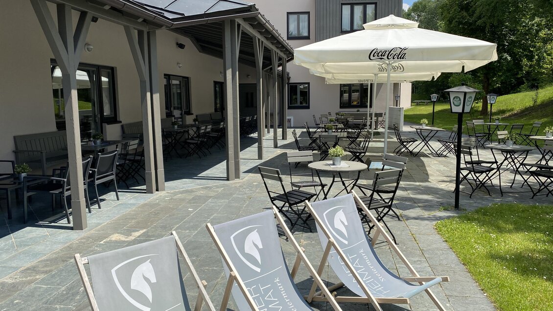 Ein einladender Außenbereich mit Liegestühlen und schattigen Tischen. Im Hintergrund befindet sich ein modernes Gebäude und ein sonniger Garten.