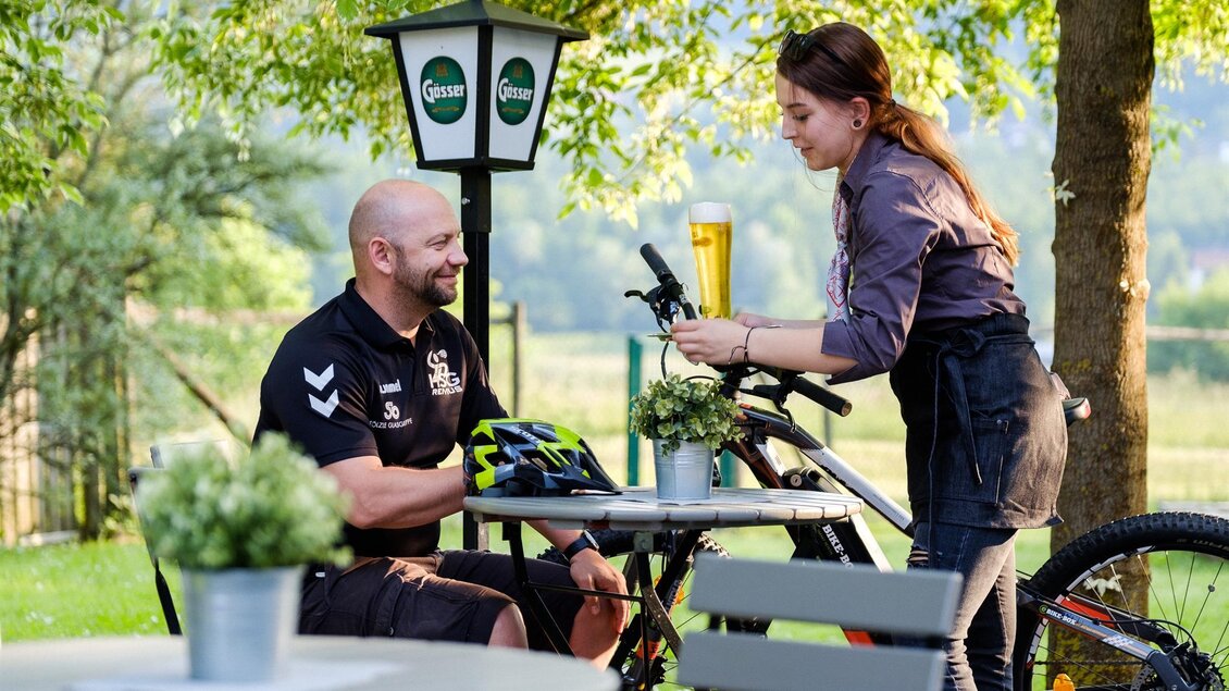 Ein Mann sitzt an einem Tisch im Freien und genießt ein Getränk. Eine Kellnerin serviert ihm in einer freundlichen Umgebung mit Bäumen und Fahrrädern. | © IQ FotoThomas Leibetseder
