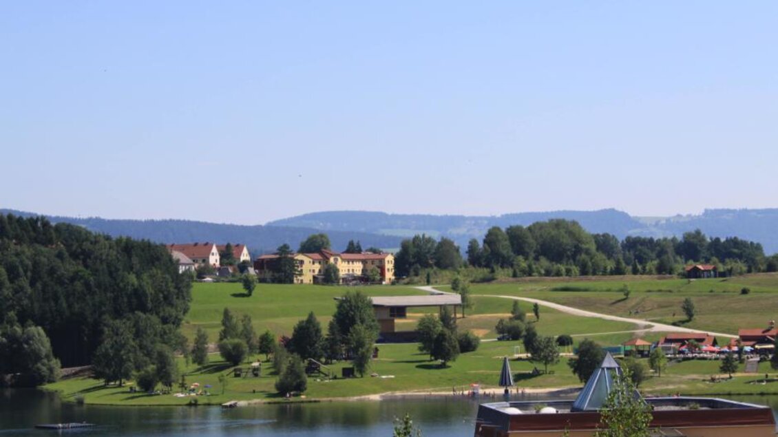 Eine malerische Landschaft mit einem ruhigen See und grünen Wiesen. Im Hintergrund sind Häuser und sanfte Hügel zu sehen. | © JUFA