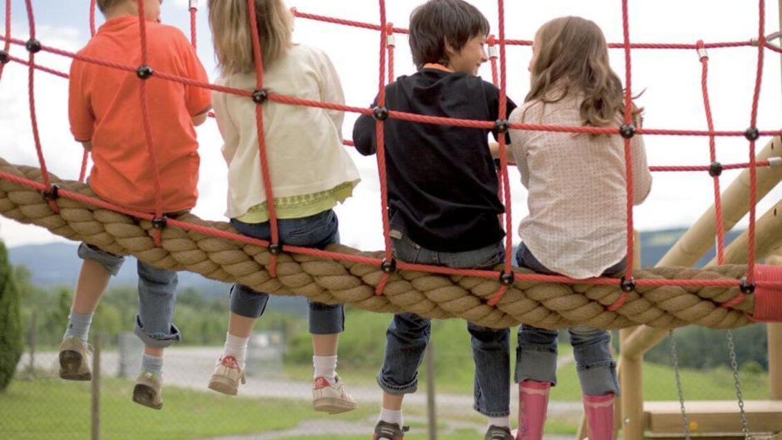 Vier Kinder sitzen auf einer Spielplatzschaukel und schauen in die Landschaft. Sie tragen unterschiedliche Kleidung und genießen die frische Luft. | © JUFA