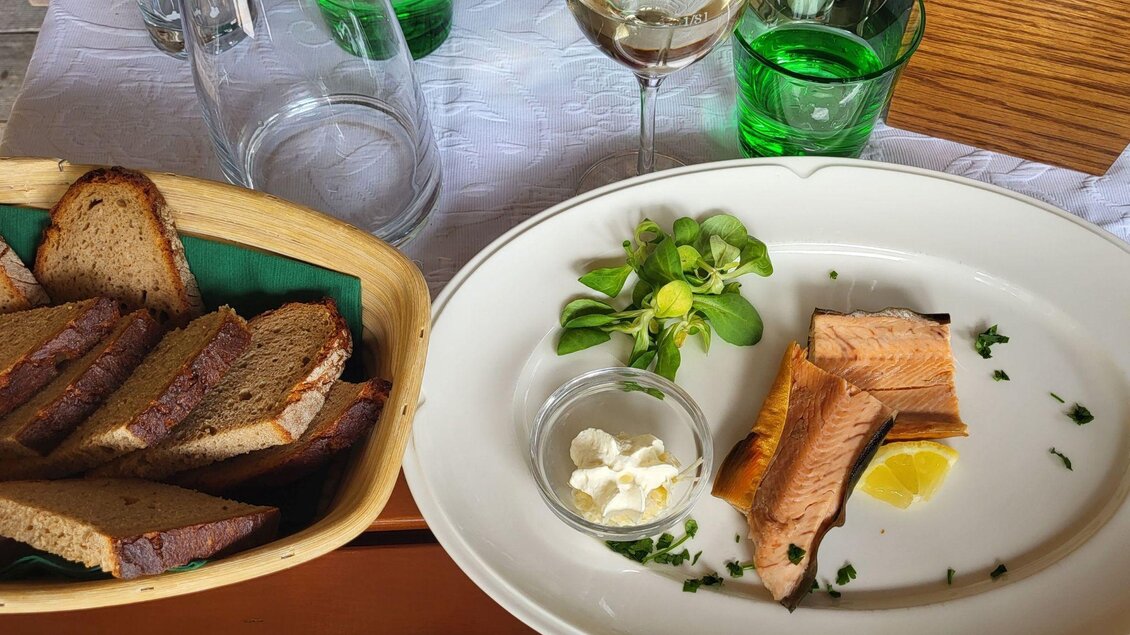 Ein Teller mit gebratenem Fisch, frischen Kräutern und einer Zitronenscheibe. Daneben befindet sich ein Korb mit Brot und einige Gläser. | © Petra Kirchschlager