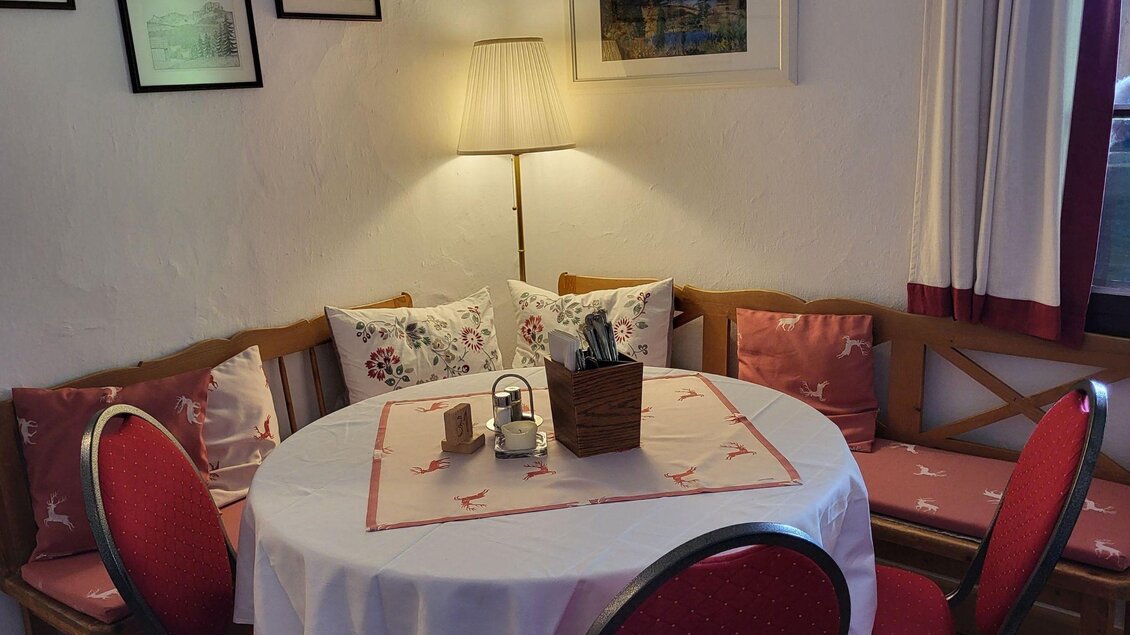 Ein gemütlicher Essbereich mit einem runden Tisch, roten Stühlen und beleuchtet von einer Lampe. Die Wände sind mit Bildern dekoriert, und es gibt Kissen auf der Bank. | © Petra Kirchschlage