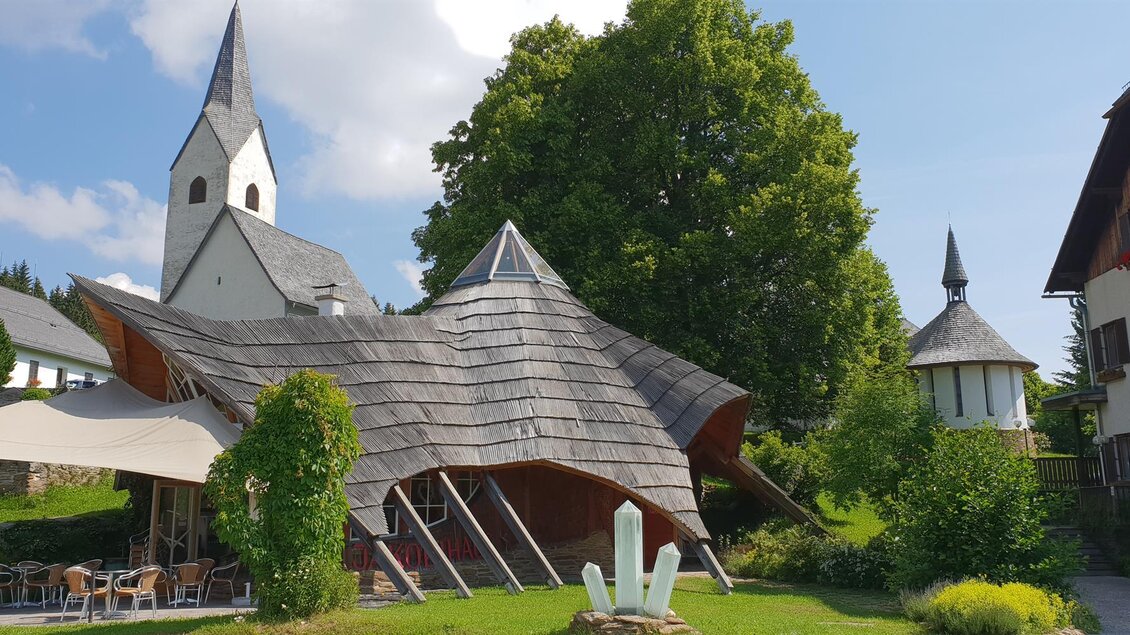 Eine moderne Architektur mit einer geschwungenen Dachform steht neben einer traditionellen Kirche. Umgeben von Bäumen und einem grünen Garten wirkt die Szene einladend und harmonisch. | © Jakobihaus Soboth