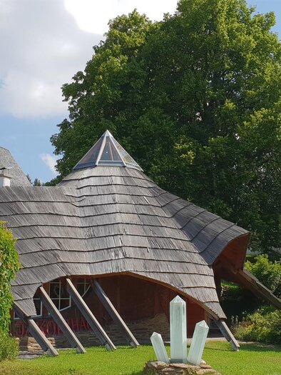 A modern architecture with a curved roof shape stands next to a traditional church. Surrounded by trees and a green garden, the scene appears inviting and harmonious. | © Jakobihaus Soboth