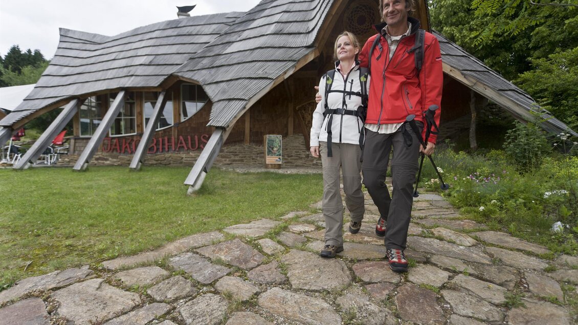 Zwei Wanderer gehen auf einem Steig vor einem interessanten, architektonisch gestalteten Gebäude. Umgeben von grüner Landschaft wirken sie entspannt und glücklich. | © Jakobihaus Soboth